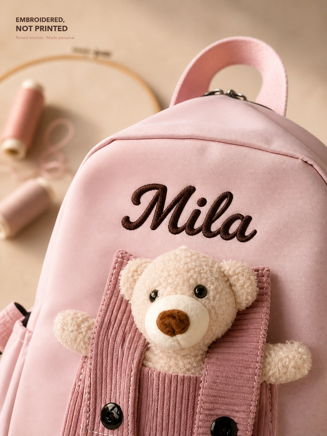 Close-up of raised Mila name embroidery above the teddy bear pocket on a pink backpack