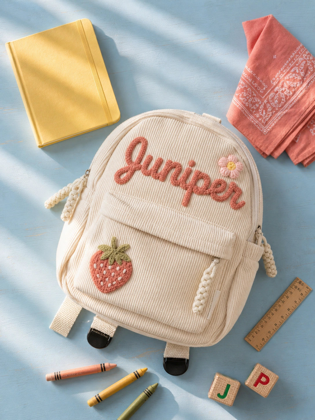 Mochila blanca de pana con nombre bordado y parche de fresa junto a material escolar