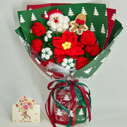 Vista frontale di un bouquet natalizio all’uncinetto con stella di Natale rossa, rose rosse, fiori di cotone, Babbo Natale e omino di pan di zenzero, accanto a un biglietto con la scritta All the best