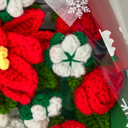 Primo piano di un fiore di cotone bianco all’uncinetto con bordo verde accanto a rose rosse e petali di stella di Natale