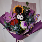 Angled view of crochet KM doll among sunflower, purple lily, red rose, tulip, lily of the valley, and green leaves