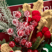 Primo piano di bacche rosse brinate, rametti sempreverdi e renna beige all’uncinetto nel bouquet natalizio