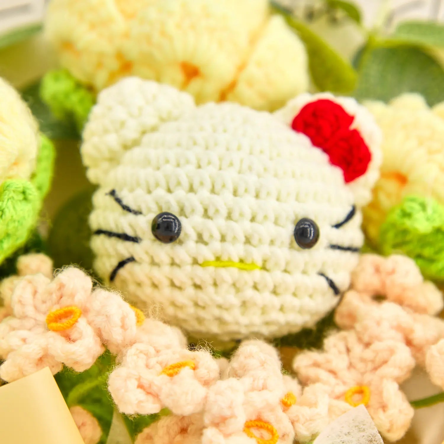 淡い黄色のバラとピーチ色の花に囲まれた、赤いリボン付きの白いかぎ針編みHKキャラクターの顔の接写