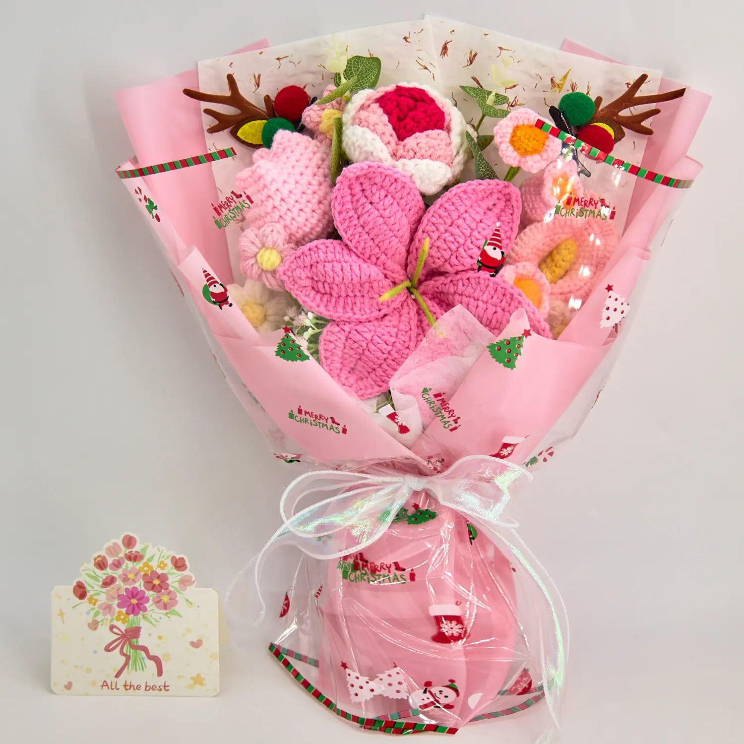 Rosa Häkelblumenstrauß mit braunen Geweihdekorationen, weihnachtlich bedrucktem Papier und einer durchscheinenden Schleife neben einer Grußkarte