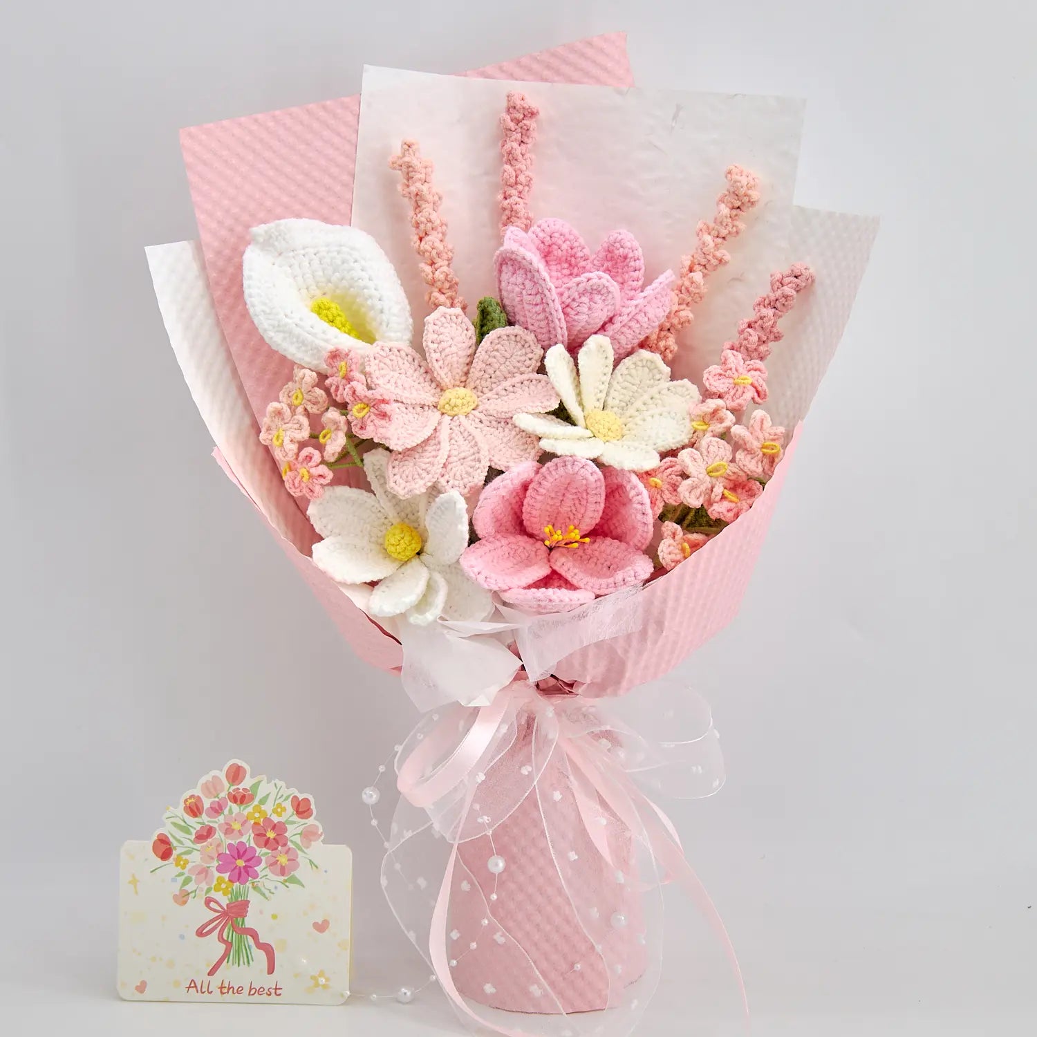 Vista frontale di un bouquet di fiori all’uncinetto rosa e bianco con incarto rosa, nastro, fili di perle e biglietto