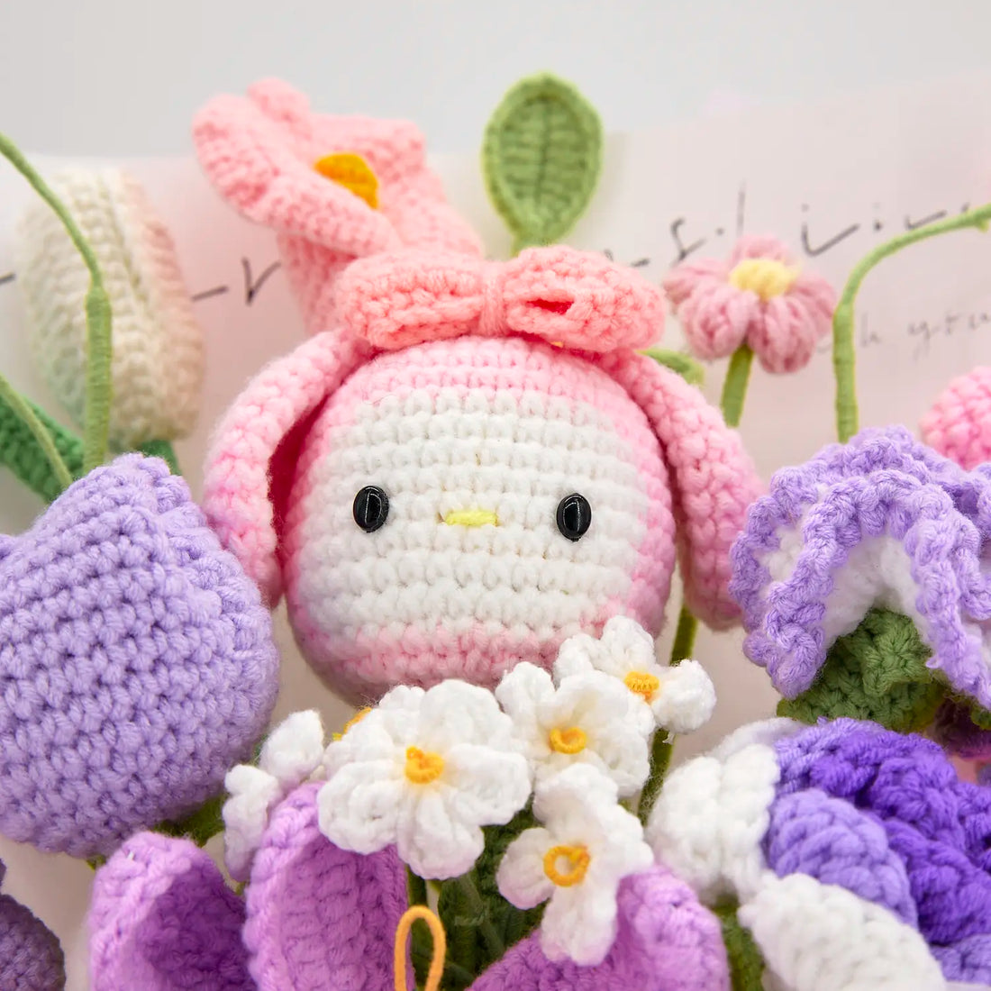 長い耳とリボンが付いたピンクのフードをかぶる白い顔のMLうさぎドールと、周囲のパープルと白の花のアップ